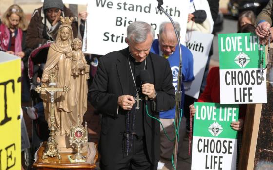 Bishop Strickland holds a rosary. People around him are bowing their heads. A person behind him holds a sign that reads: "We stand with Bishop Strickland."