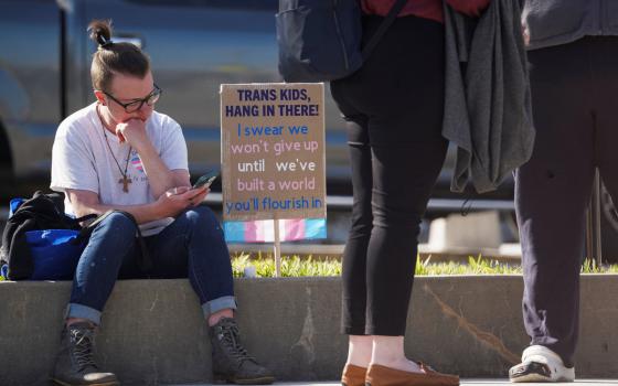 Person sits on sidewalk next to a sign that says, "Trans kids, hang in there." 