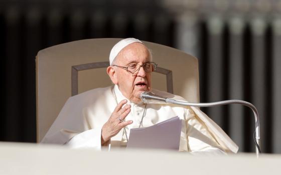 Pope Francis speaks into a microphone while sitting in a white chair and holding a paper