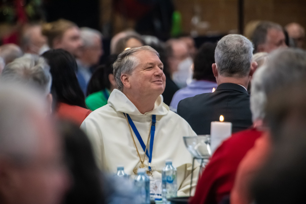 An older white man wearing a white robe and pectoral cross sits in a crowded room