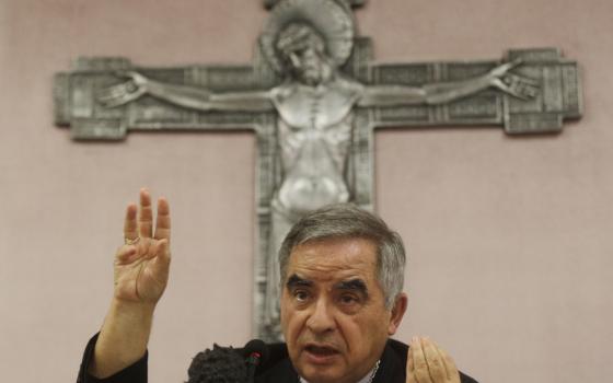 An older white man raises his hand while in front of a large silver crucifix on the wall